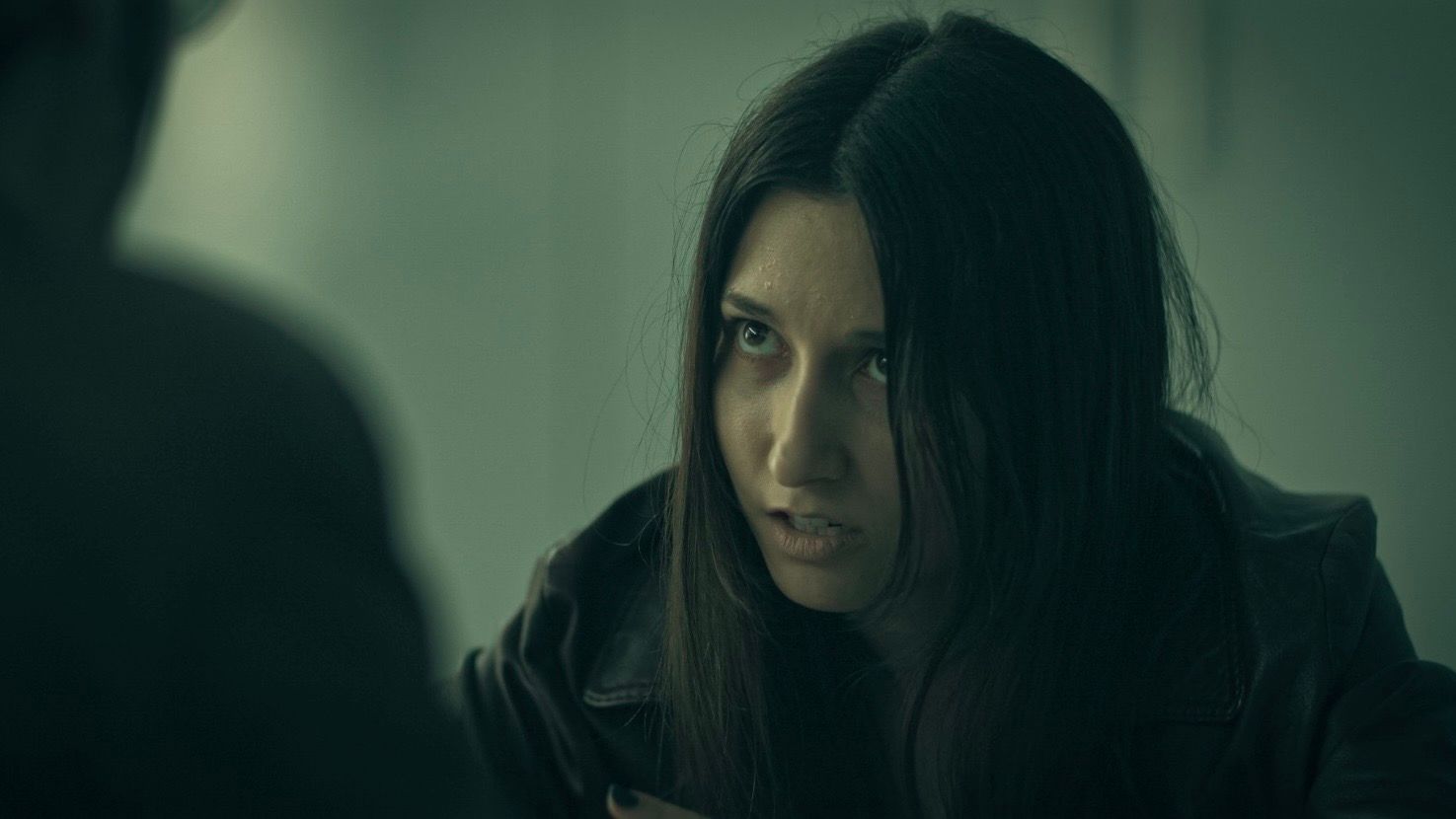 Jessica Curado in character, leaning forward in a leather jacket and looking intently at someone opposite her.